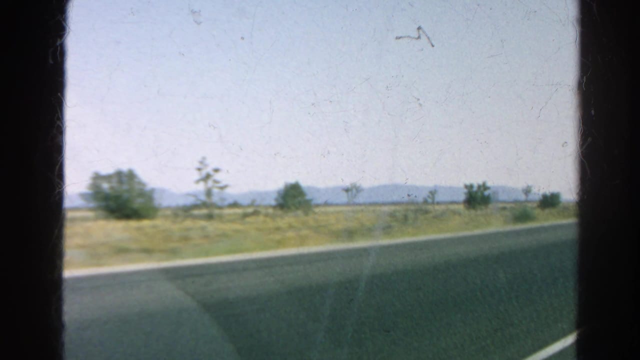 1966 California home-movie frame of a desert road seen from a moving car, with shrubs, Joshua trees, and low mountains in the distance.