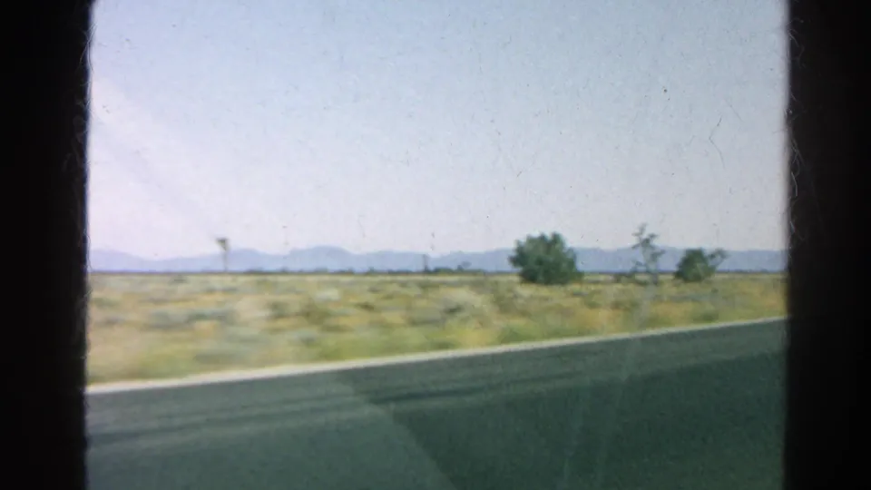 1966 8mm frame from a moving vehicle, showing blurred California desert roadside vegetation, Joshua trees, and a distant mountain line.