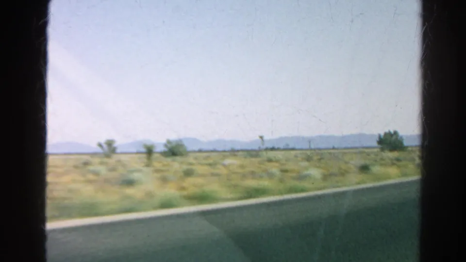 1966 California road-trip view from a moving vehicle, with desert vegetation, Joshua trees, a paved road edge, and hazy mountains.