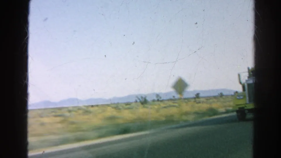 The yellow tanker truck drops back at the frame edge as the moving car continues along the desert highway.