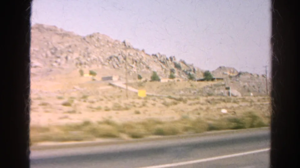 1966 8mm view of a rocky California desert roadside with scrub, highway markings, and a small yellow sign.