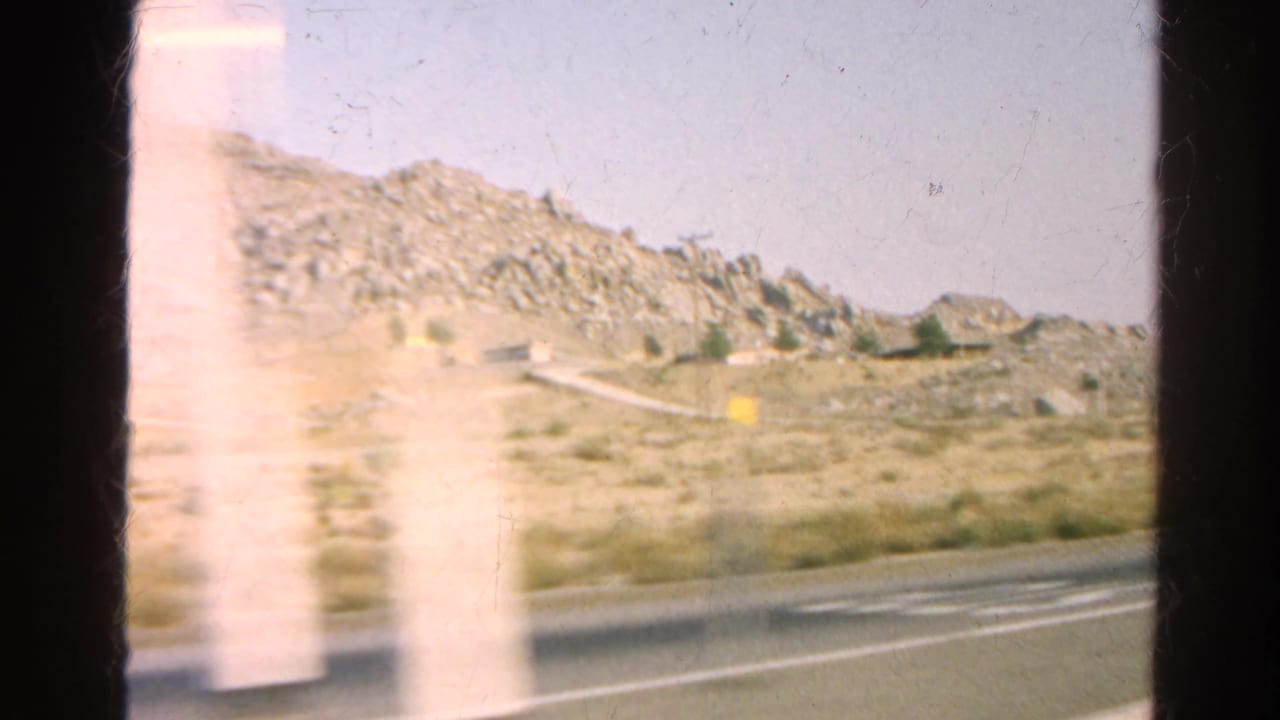 1966 8mm view of a dry California roadside with rocky hills, sparse scrub, and dark car-window edges.