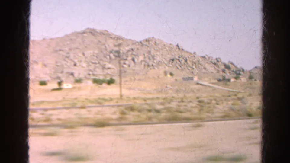 The camera looks across a roadside shoulder toward rocky hills and sparse vegetation during a 1966 California drive.