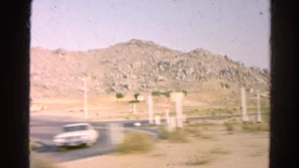 A blurred white car appears on the road near roadside posts, with dry hills and scrub beyond in this 1966 8mm frame.