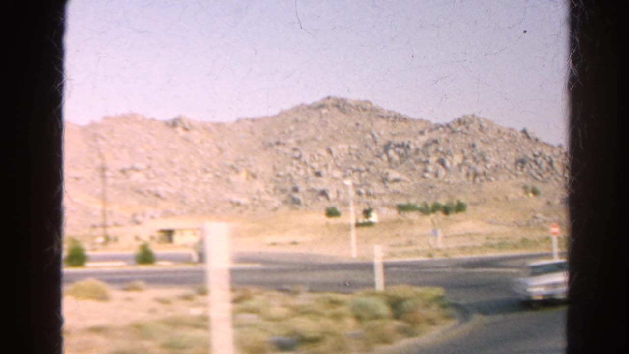 1966 8mm view from a moving vehicle of rocky California terrain, sparse scrub, road, and window-frame edges.