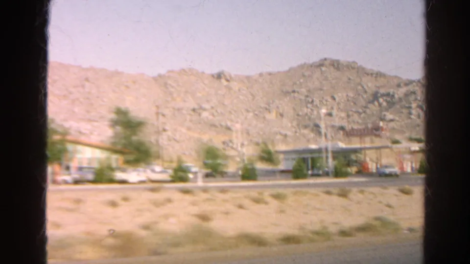 Moving 8mm view of a low roadside building, service-station area, cars, scrub, and rocky terrain in 1960s California.