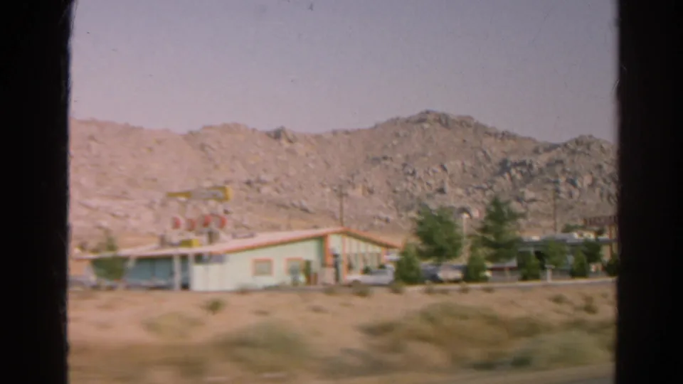 1966 8mm frame of a low roadside commercial building, unreadable sign, parked cars, scrub, and rocky hills.