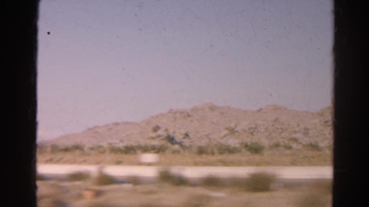 1966 8mm road-view frame of arid hills, roadside scrub, and pale sky, partially framed by a dark car window.