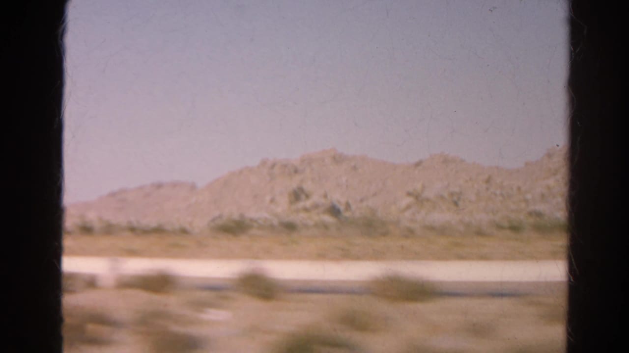 Arid hills and low roadside vegetation viewed from a moving vehicle.