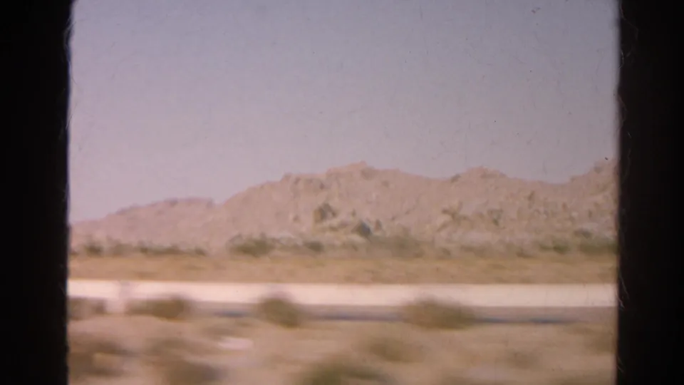 1966 California home-movie still showing low desert hills, scrub, and a bright roadside strip from inside a moving car.