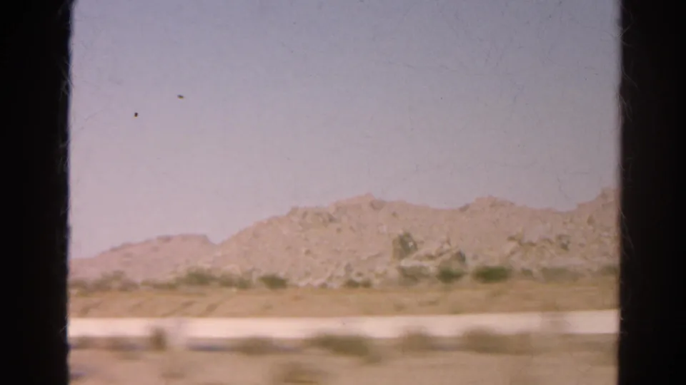 1966 8mm frame of a low desert ridge, sparse vegetation, and sandy roadside ground framed by black vehicle-window edges.