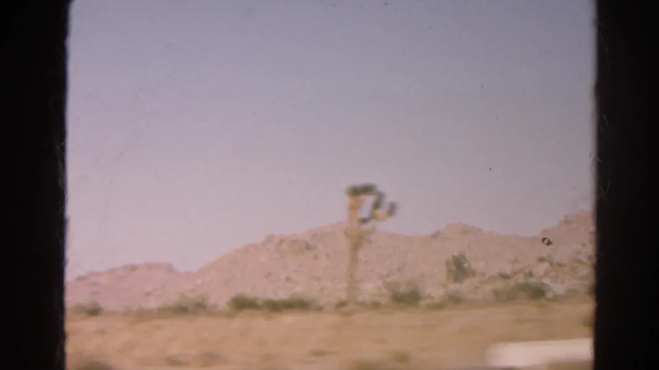 1966 8mm road-trip still showing desert hills, scattered scrub, and a small pole-mounted object near the roadside.