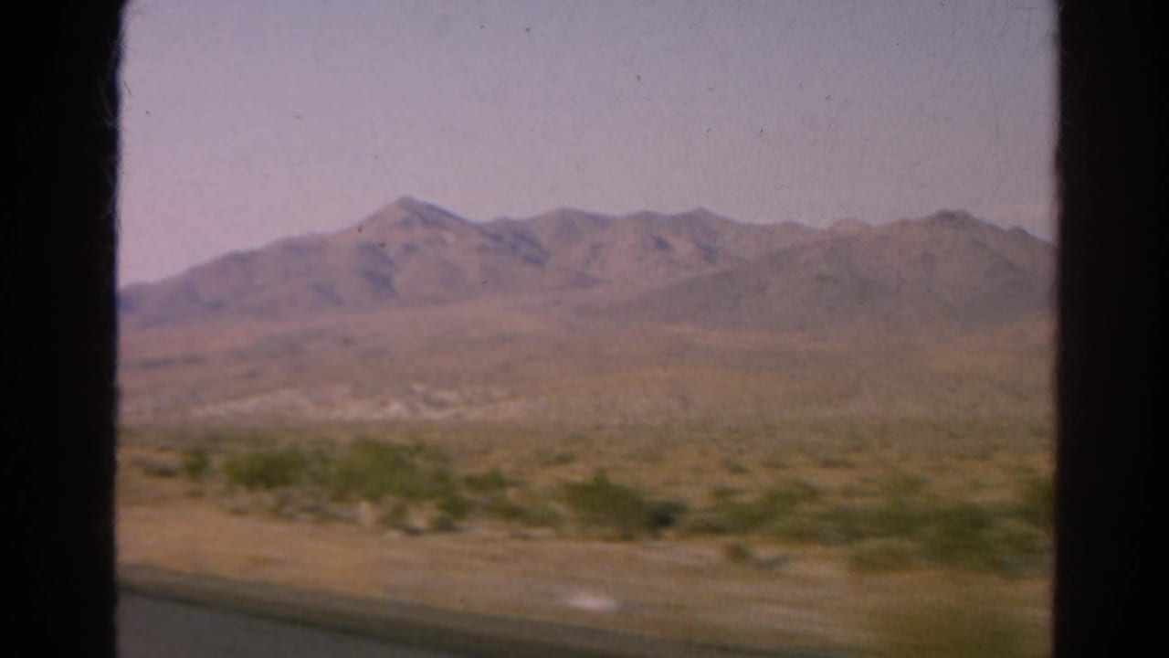 1966 8mm frame near Silver Lake, California, with distant rocky mountains, scrub, and a road shoulder from a moving vehicle.