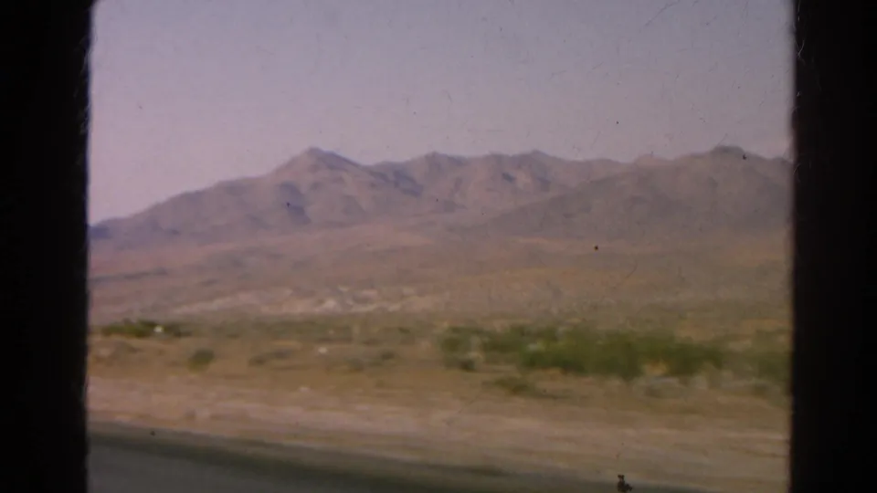 1966 8mm road-view frame near Silver Lake, California, showing desert scrub, a paved road edge, and distant mountains.