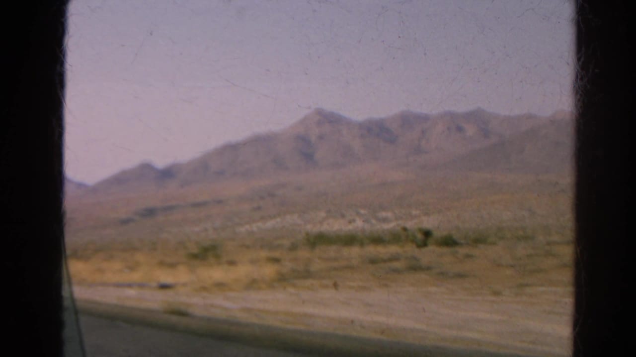 Desert road seen through a car window with distant mountains under a hazy sky.