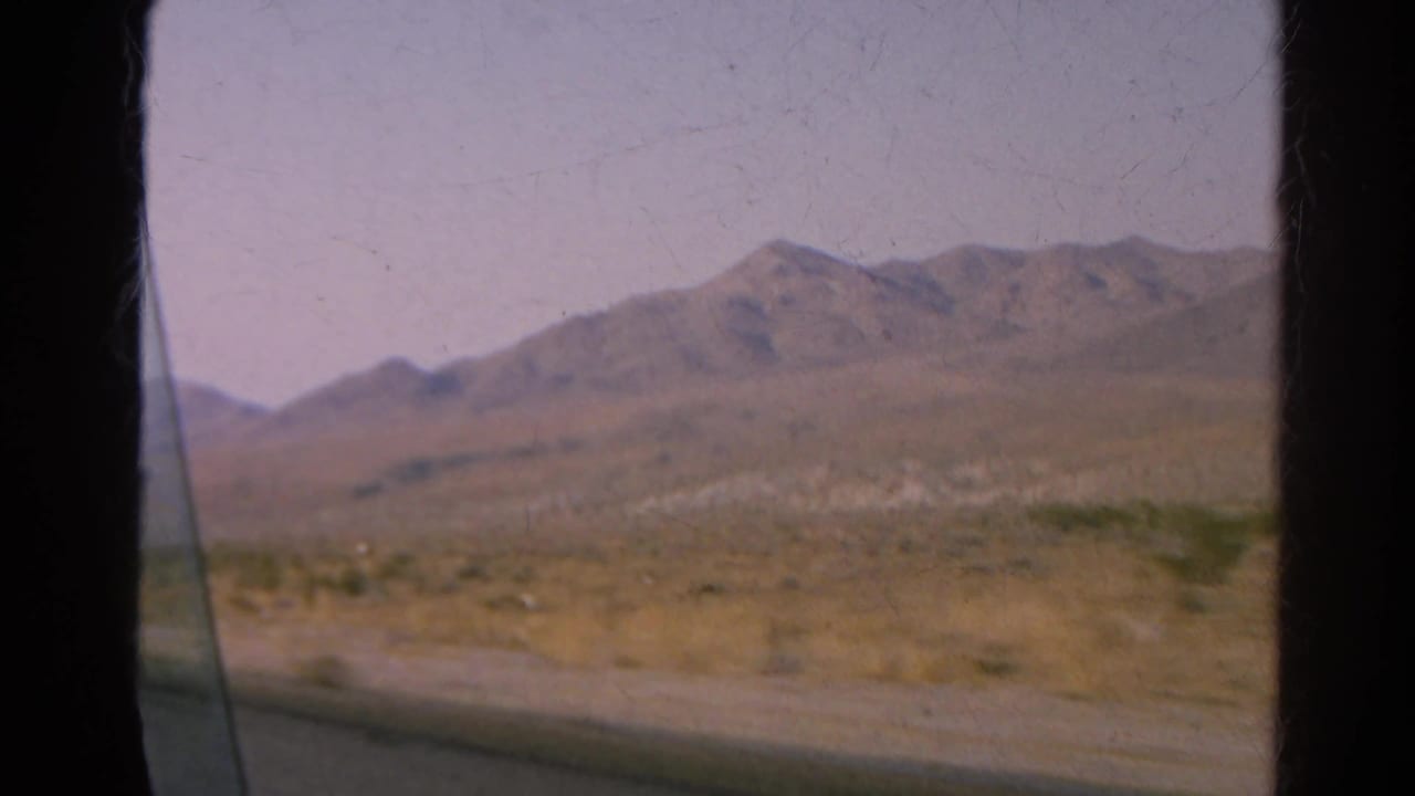 Moving-car view across a dry desert landscape with mountains, likely from the same 1966 road-trip clip.