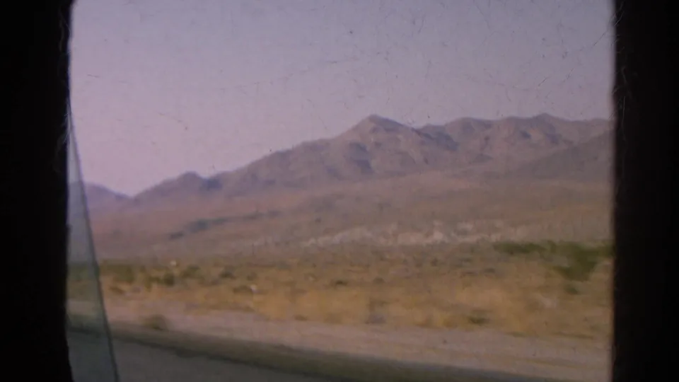 Moving-car view across a dry desert landscape with mountains, likely from the same 1966 road-trip clip.
