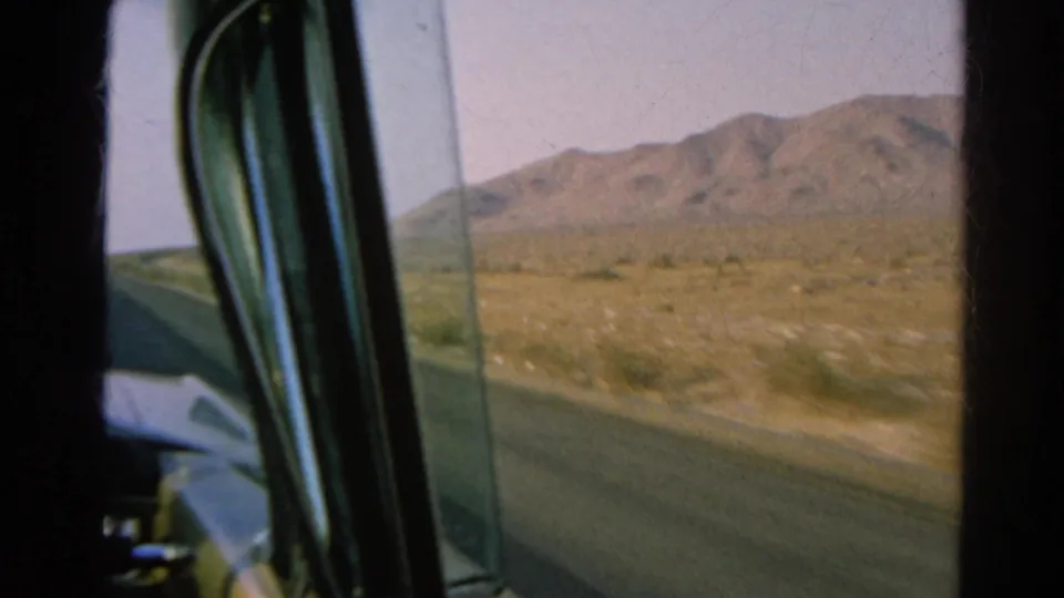 Inside-car 8mm view of a desert highway shoulder and distant hills, with part of the vehicle visible at left.