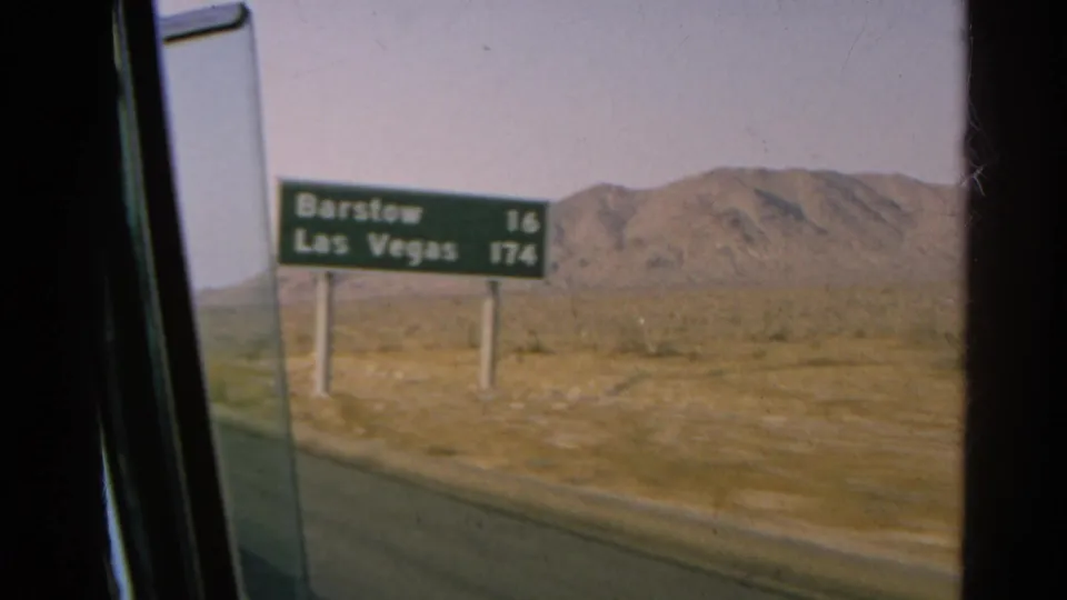 1966 8mm frame from a moving car showing a roadside sign for Barstow and Las Vegas in a desert landscape.