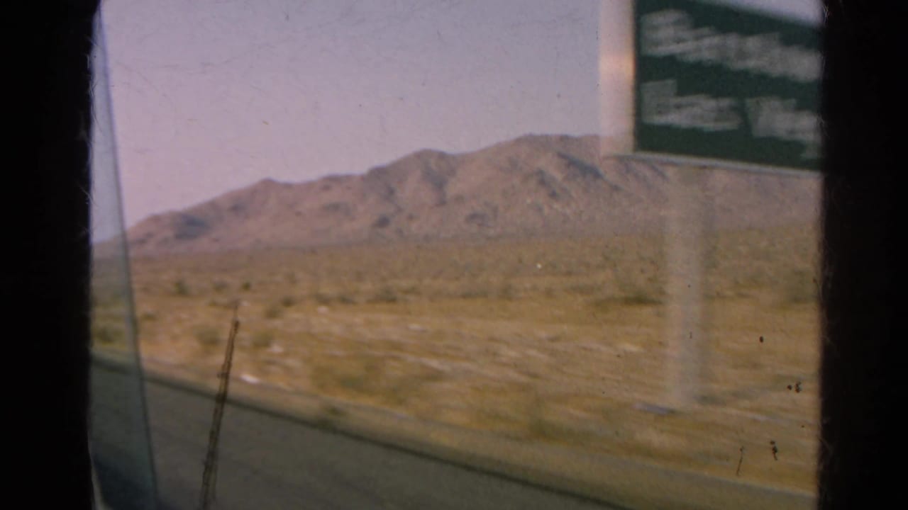 1966 8mm home-movie frame from Silver Lake, California, showing a roadside view, blurred sign, dry open land, and distant mountain.