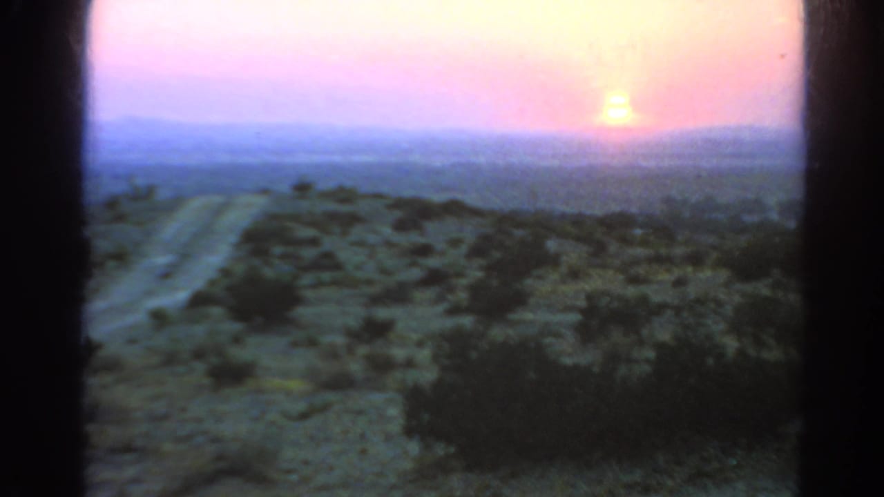 A soft 1966 8mm frame shows the sun near the horizon over scrubby California desert terrain and a lightly curving dirt road.