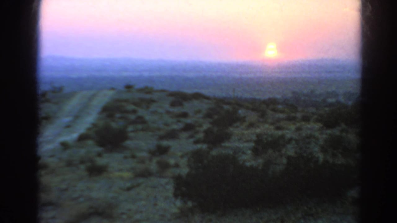 The 8mm home-movie frame captures a bright low sun, pastel sky, distant ridgeline, sparse shrubs, and a desert road in 1966 California footage.