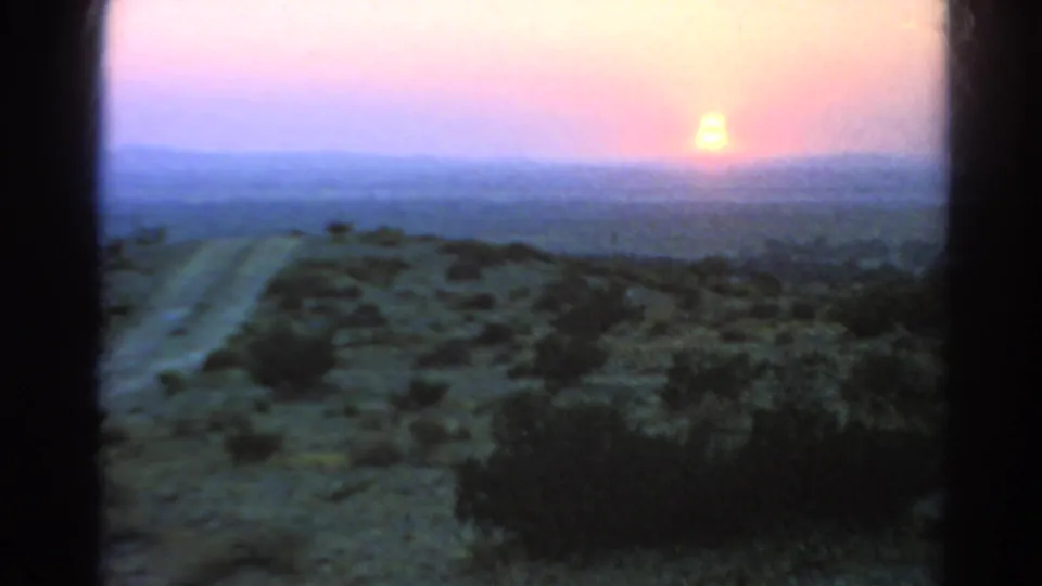 The 8mm home-movie frame captures a bright low sun, pastel sky, distant ridgeline, sparse shrubs, and a desert road in 1966 California footage.