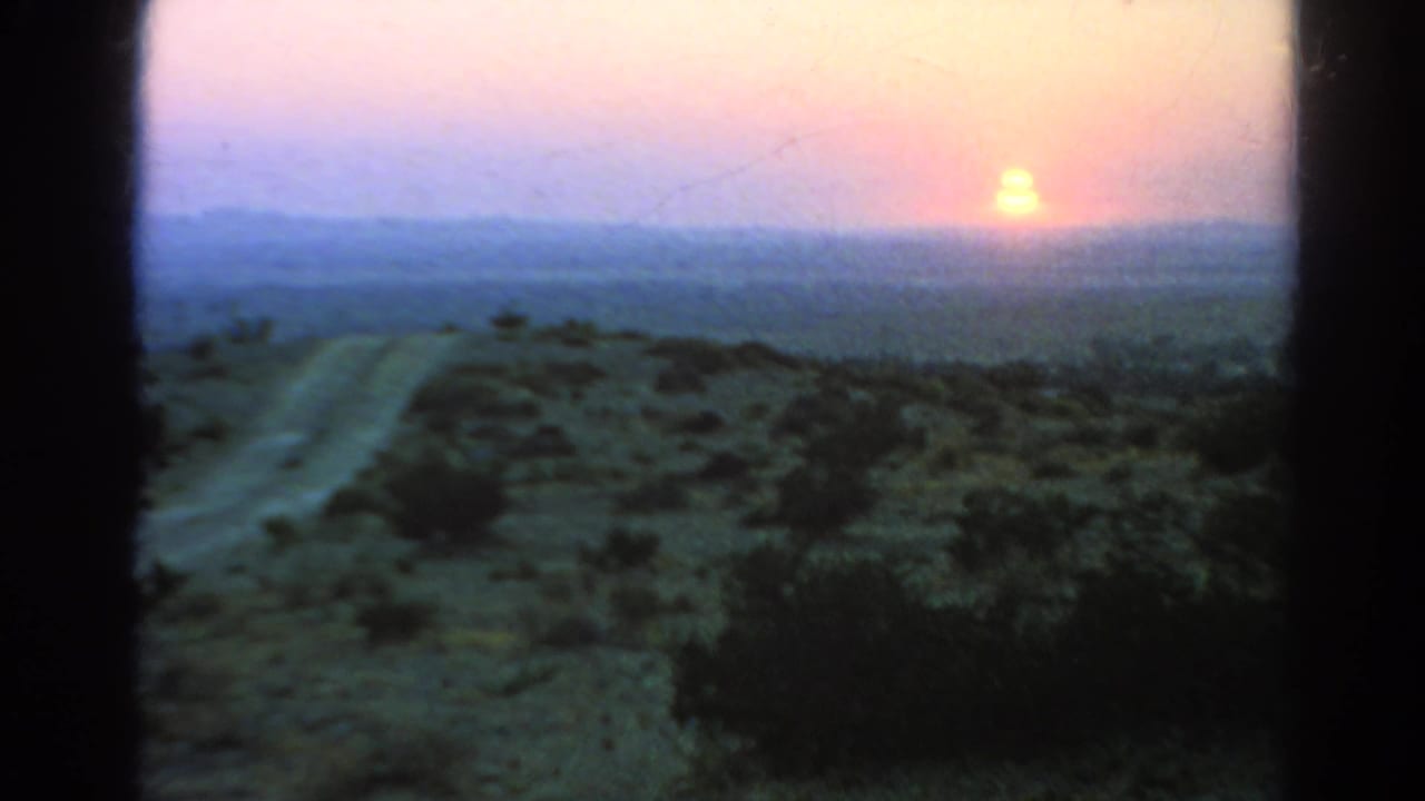 A grainy 8mm view shows a desert road, dark foreground brush, and the setting sun against a pink-yellow sky near Silver Lake, California.