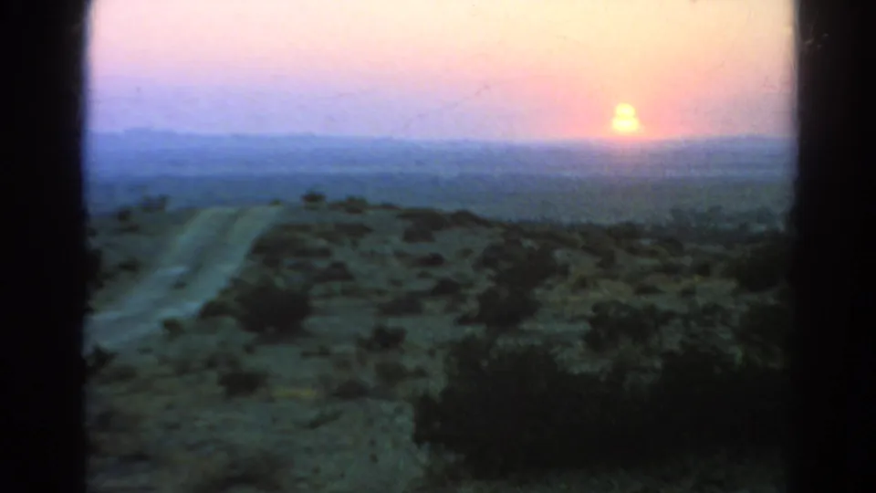 A grainy 8mm view shows a desert road, dark foreground brush, and the setting sun against a pink-yellow sky near Silver Lake, California.