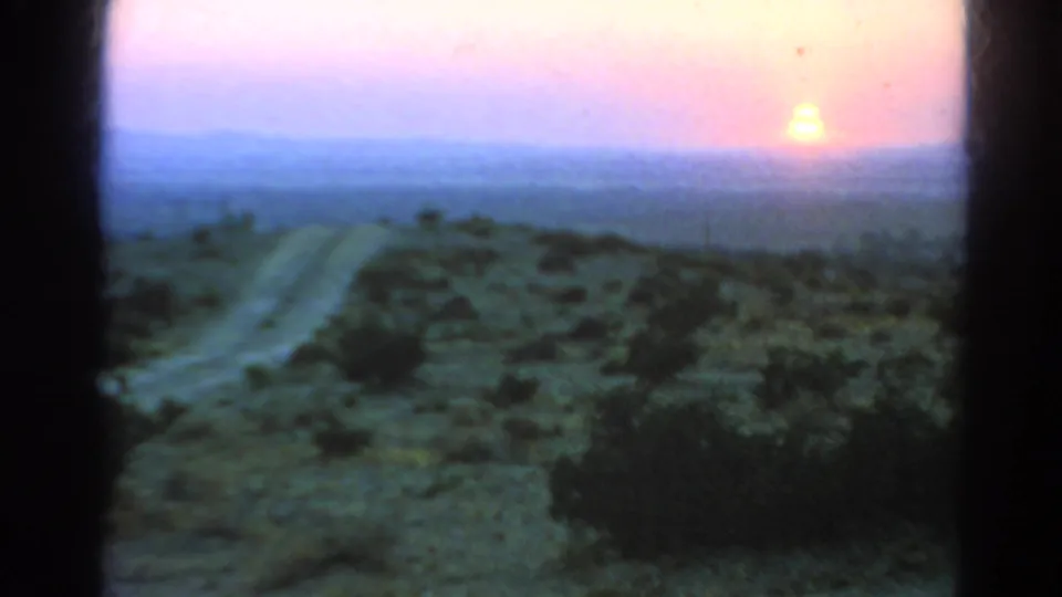A soft 8mm frame shows a pale dirt track through scrubland under a pink and blue sunset sky, with dark film borders visible.