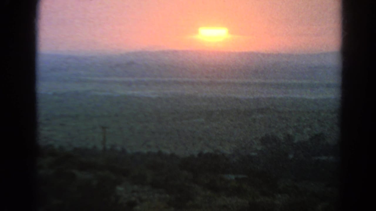 The 8mm frame centers on the orange sun over a hazy arid horizon, with dark scrub and soft color shifts across the lower frame.