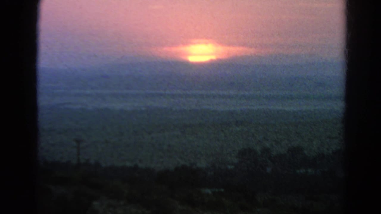A soft 1966 8mm sunset view shows the sun partly diffused by cloud over a broad arid landscape, with dark foreground scrub and frame edges.