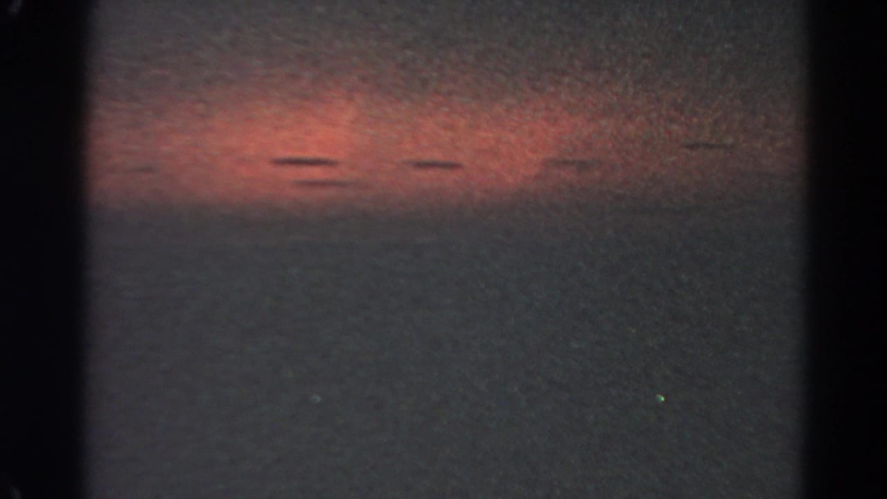 Underexposed opening frame from the 8mm clip, with mostly dark film area and a faint reddish band of light.