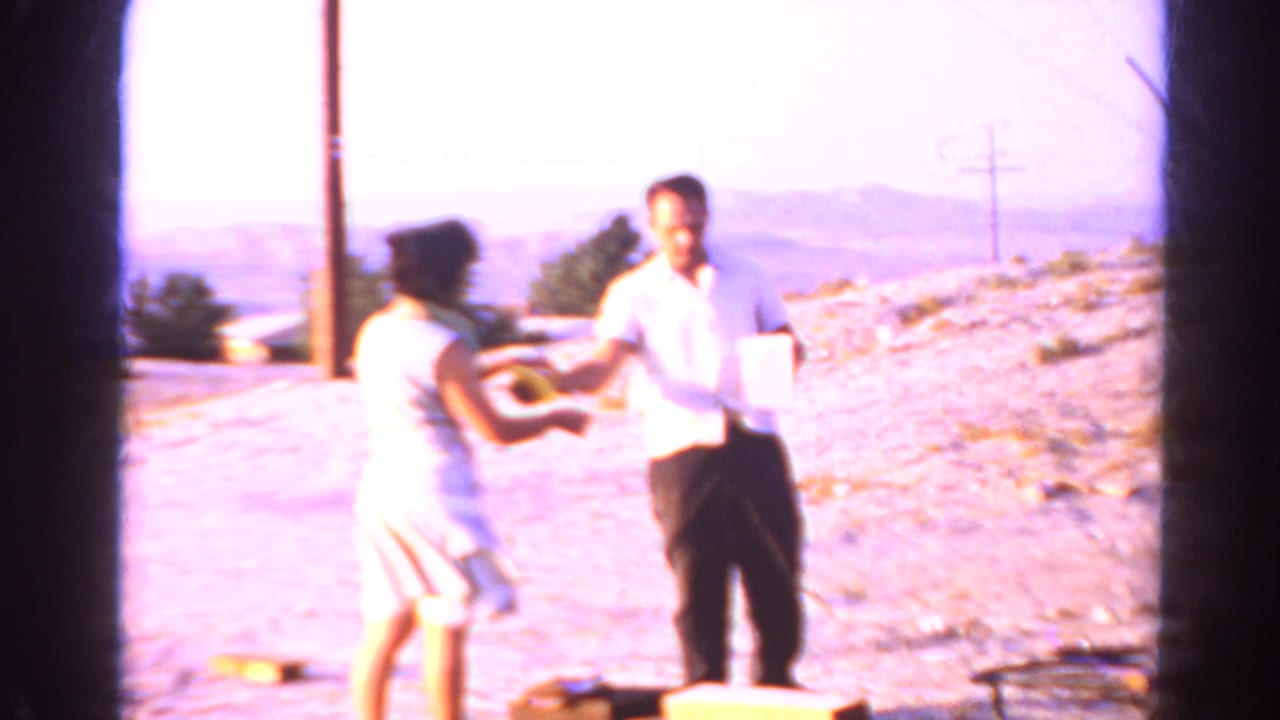A woman outdoors appears to hand a pitcher toward a man on a dirt hillside.