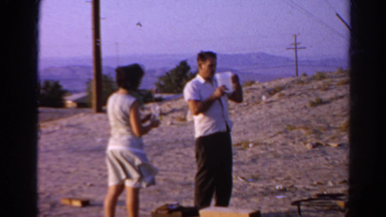 A woman in a sleeveless dress stands beside a man in a white shirt holding a pitcher, with utility poles and distant hills behind them.
