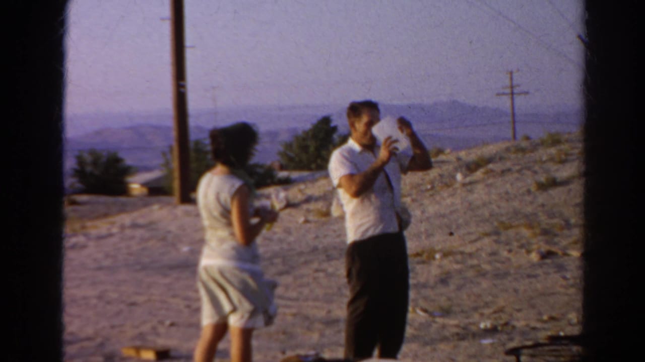 Woman faces a man holding a jug on open dirt ground with hills in the background.
