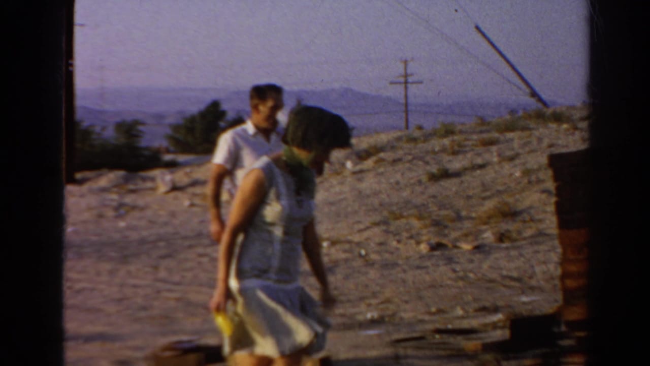 The woman bends near the ground with a yellow object while the man stands behind her at the dusty hillside location.