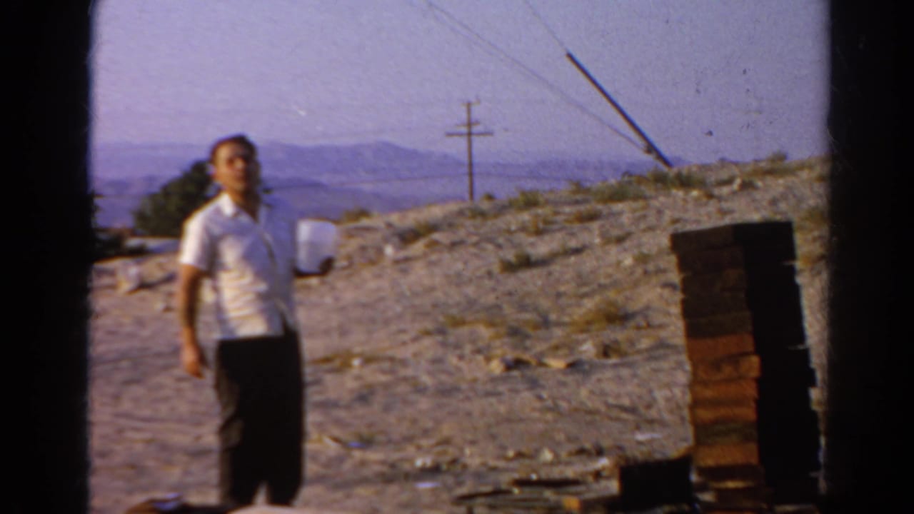 Man in a white shirt stands on a dusty hillside near stacked bricks and distant hills.