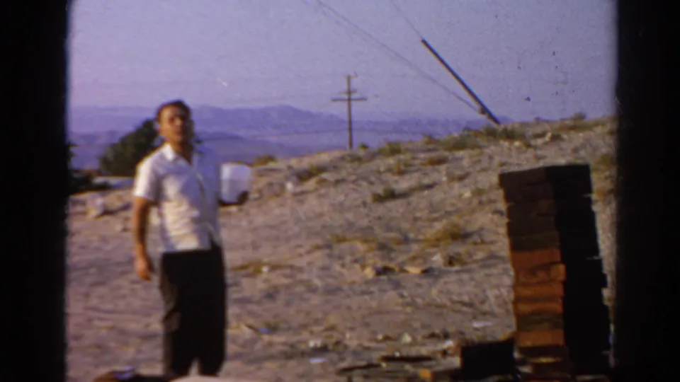 Adult man in a white shirt stands outdoors in Silver Lake, California, near stacked bricks, a dusty slope, utility pole, and distant hills.