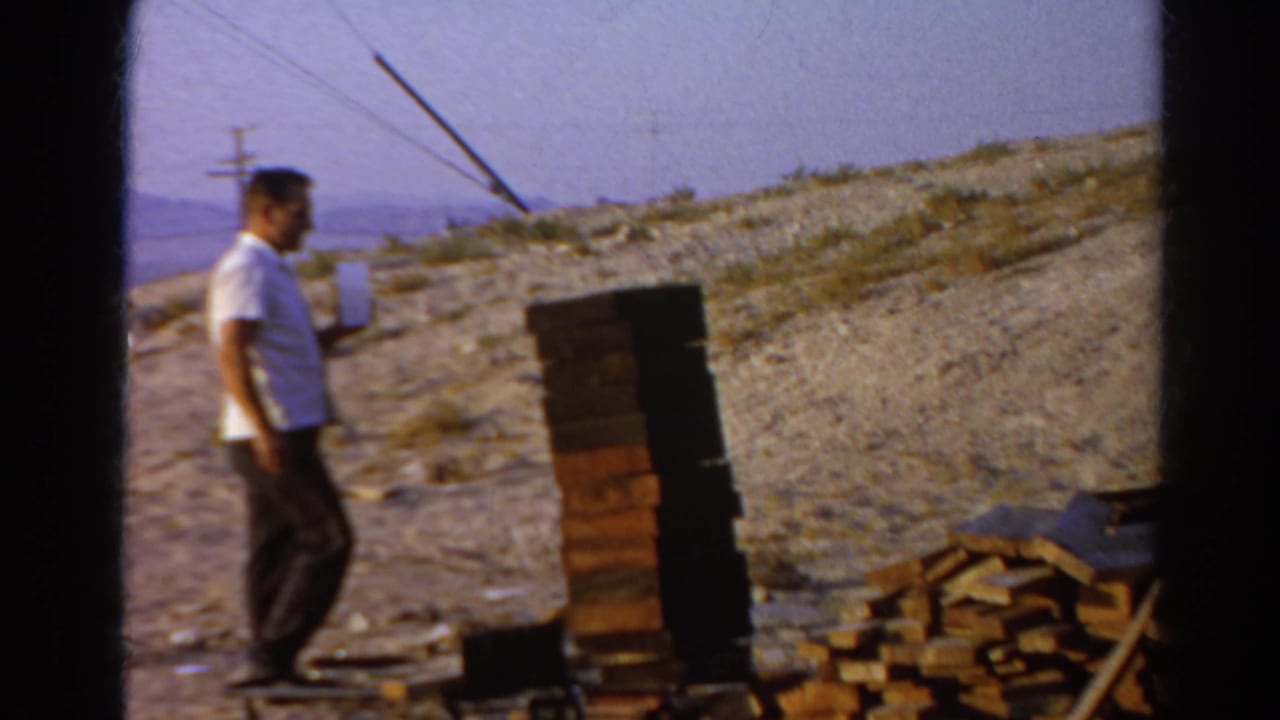 Adult man in a white shirt moves toward stacked bricks and scattered wood on a dry Silver Lake hillside.