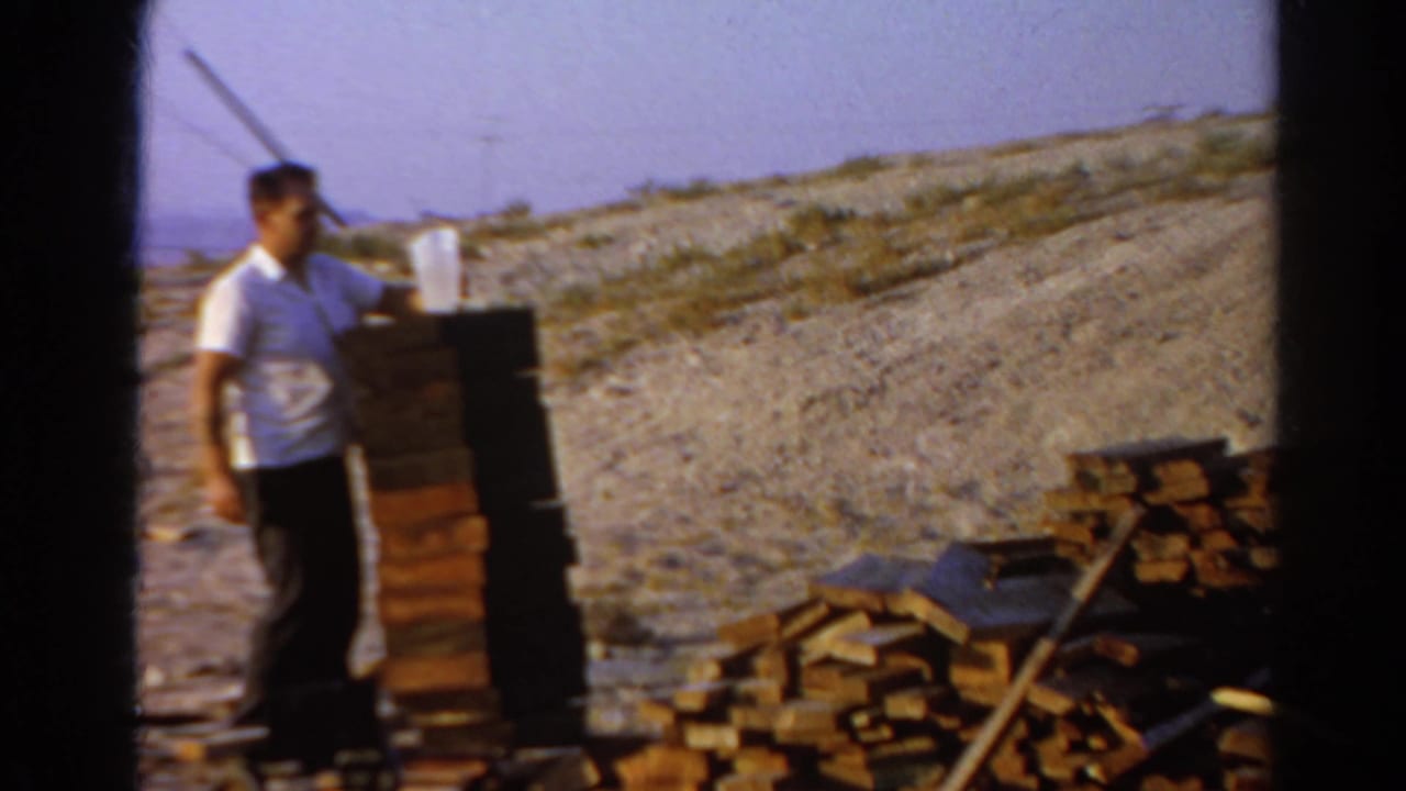 Adult man on a Silver Lake hillside appears to set or hold a white cup by a stacked brick column, with scattered boards in the foreground.