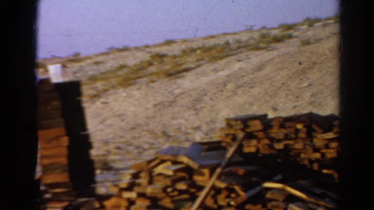 1966 Silver Lake home-movie frame showing stacked lumber, scrap boards, and dry sloped ground under a pale sky.