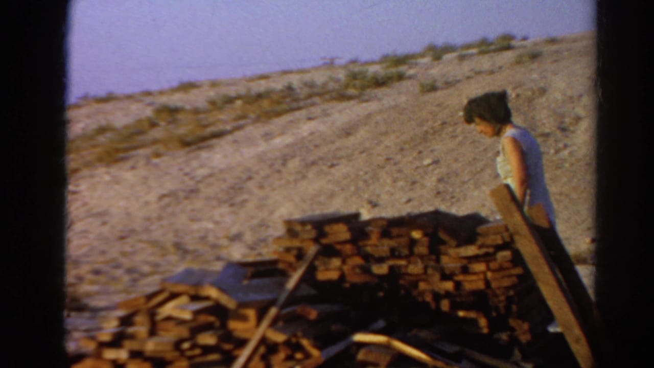 Woman in a light sleeveless dress stands beside stacked lumber on dusty ground.