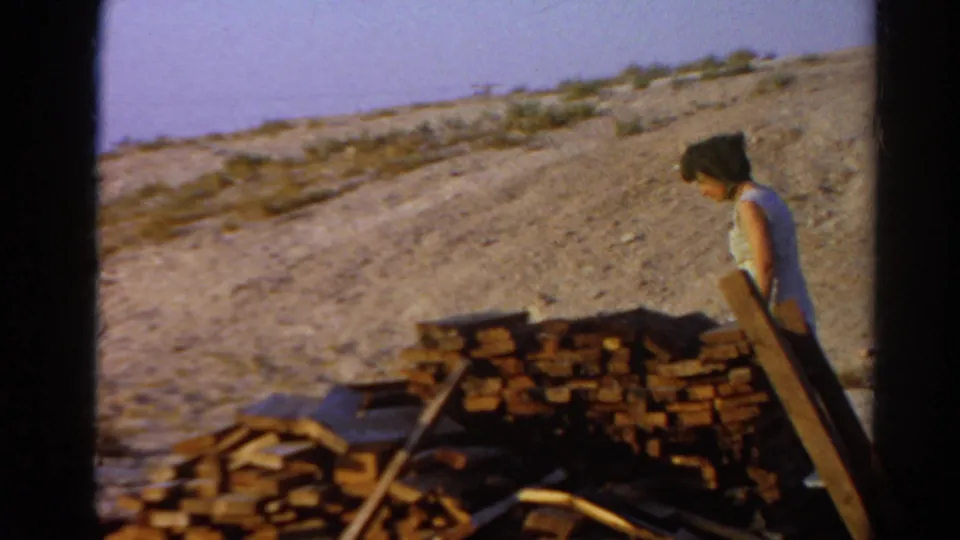 1966 Silver Lake frame with a woman beside wood stacks in a dry outdoor construction area.