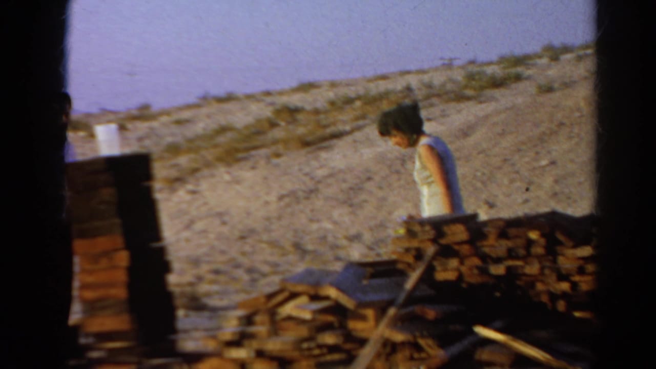 1966 Silver Lake home-movie frame showing a woman moving past lumber piles at an outdoor construction site.