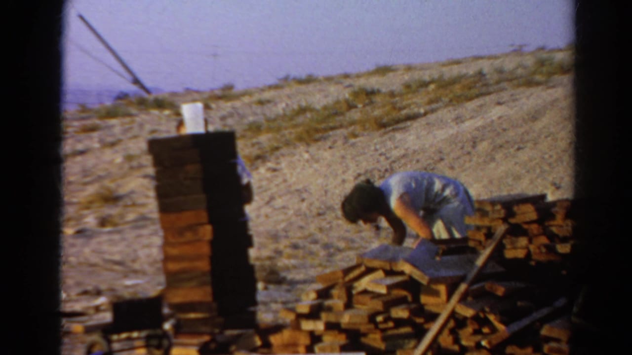 In Silver Lake, California, 1966, a man bends over beside bricks and wood planks at an outdoor construction site.