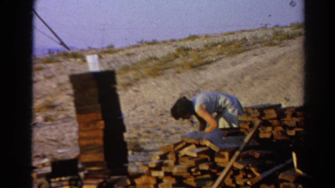 Man bends near a brick stack and lumber with a dry hillside behind him.