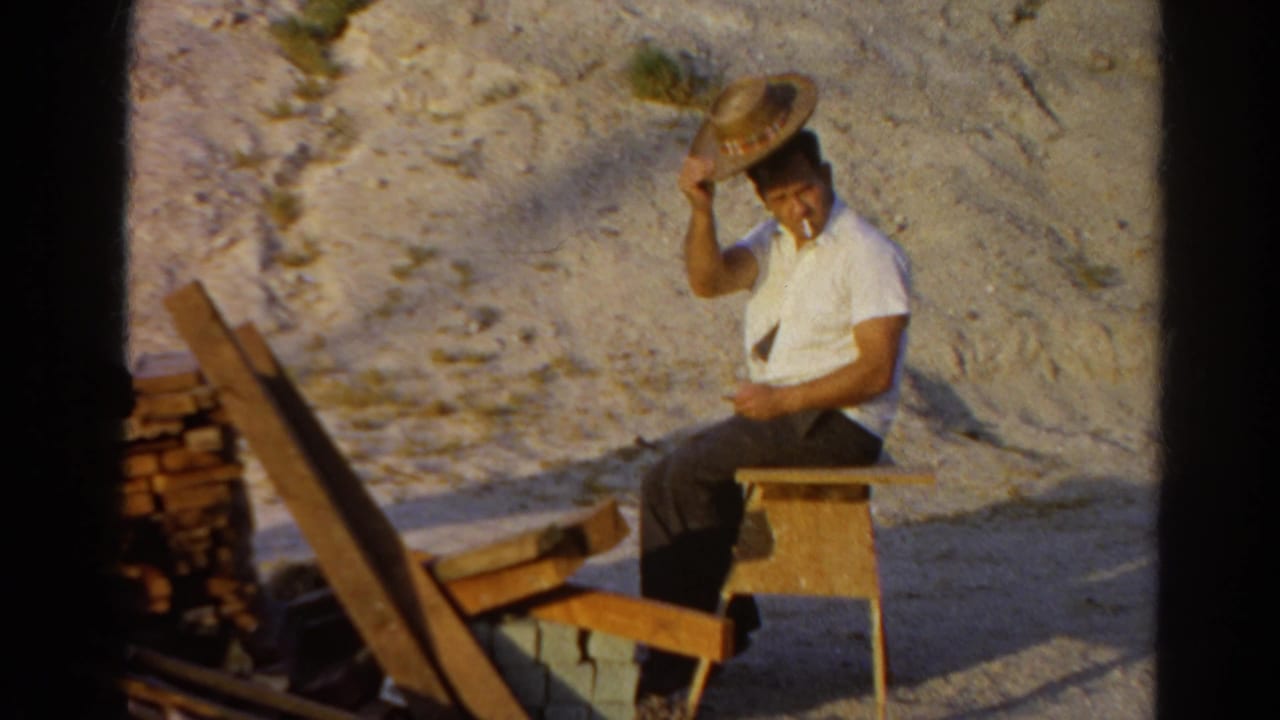 A seated man in a white shirt adjusts a straw hat while smoking near lumber and bricks in Silver Lake, 1966.