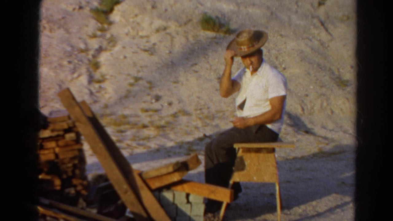 The man sits on a simple wood chair, holding his straw hat and smoking beside bricks and planks in Silver Lake, 1966.