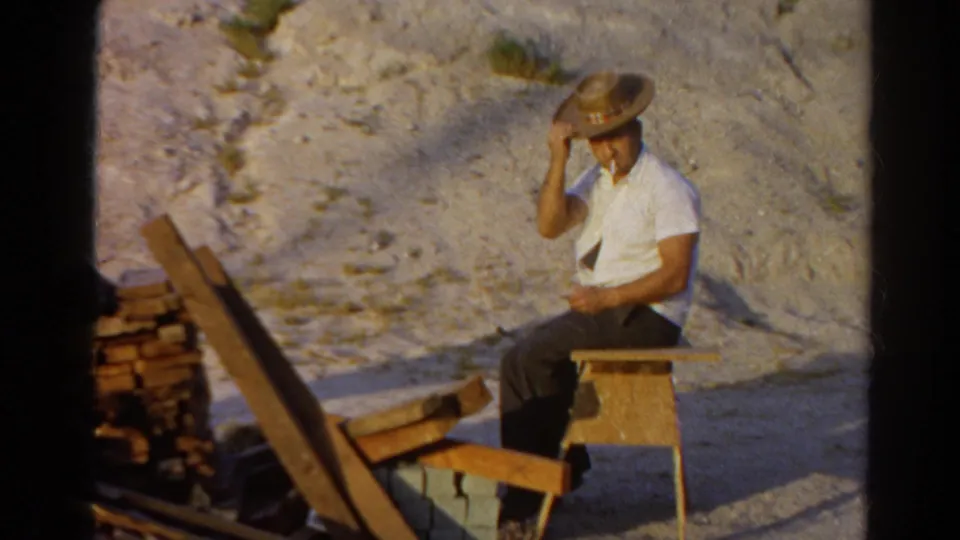 The man sits on a simple wood chair, holding his straw hat and smoking beside bricks and planks in Silver Lake, 1966.